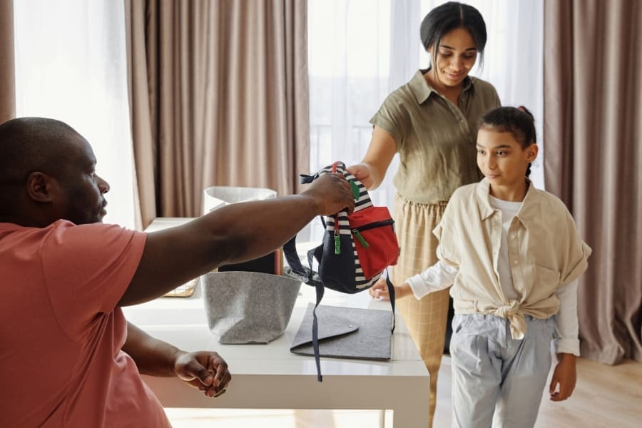 A man hands a colorful backpack to a girl, making school runs less stressful, while a woman stands beside her in a room with curtains and a table.