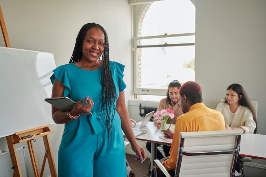 A confident working mother in a blue outfit stands holding a tablet, smiling at the camera, while three people sit at a table having a discussion in a bright office, reflecting confidence in leadership.