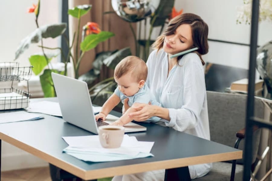 A working mom multitasks at her desk on a laptop, holding her baby and talking on the phone, with plants and office items in the background—a true example of balancing work and self-care.