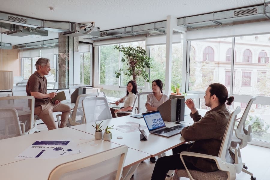 Four people sit around a table in a modern office, engaged in a discussion about network infrastructure planning. One person stands and talks, while another uses a laptop. Large windows provide natural light.