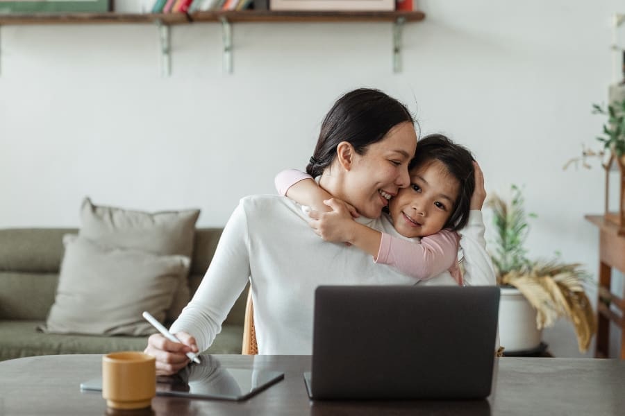 A woman works at a table with a laptop and tablet while a young child hugs her from behind, reflecting the joys and challenges of work-life balance in a home setting.