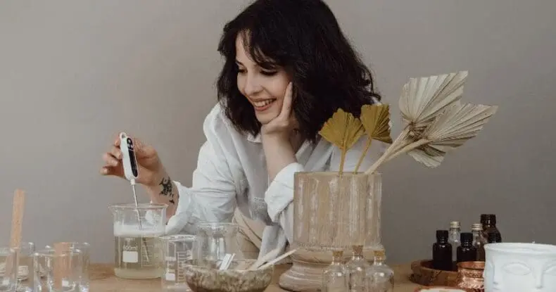 A woman in a white shirt smiles while mixing a liquid in a beaker with a thermometer, surrounded by glassware, dried leaves, and small bottles on a table.