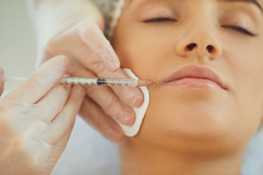 A close-up of a person receiving a cosmetic injection in the lips, showcasing family friendly healthcare careers ideal for working mums in healthcare, with gloved hands holding a syringe and a cotton pad.
