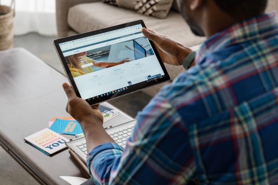 A person in a plaid shirt uses a tablet at a table with a laptop and colorful notebooks, exploring LinkedIn hacks for an efficient 2025 job search. The tablet displays a website with a search bar and images.