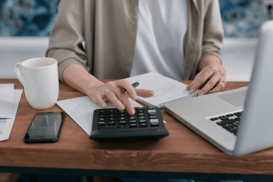 Person at a desk using a calculator next to a laptop, notebook, and coffee mug while planning long-term financial security.