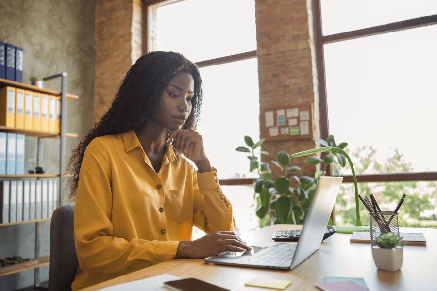 A woman in a yellow shirt sits at a desk, looking at a laptop computer whilst engaging in remote work.