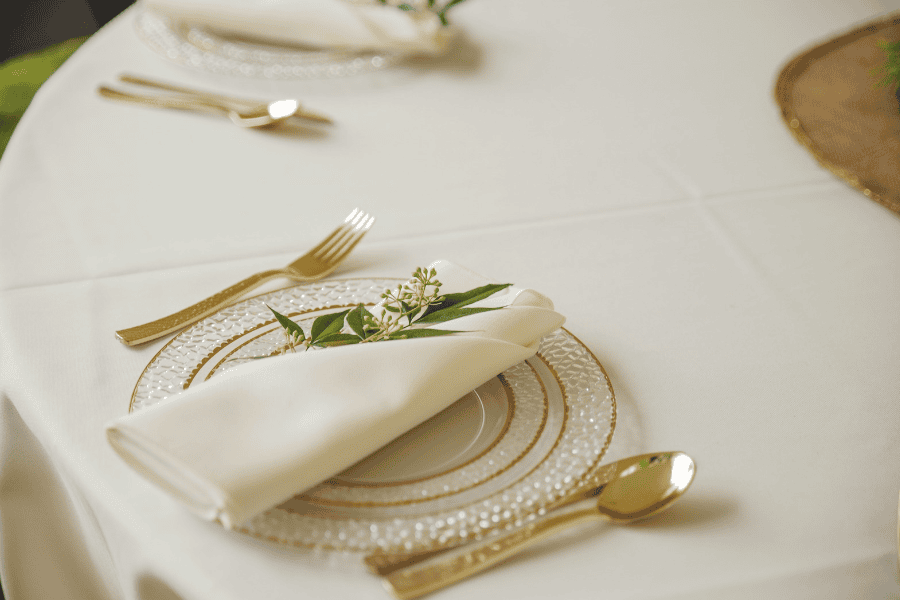 A clear, gold-rimmed plate and folded cream serviette topped with greenery sit on a white tablecloth at this event table setting, finished with gold-toned cutlery.