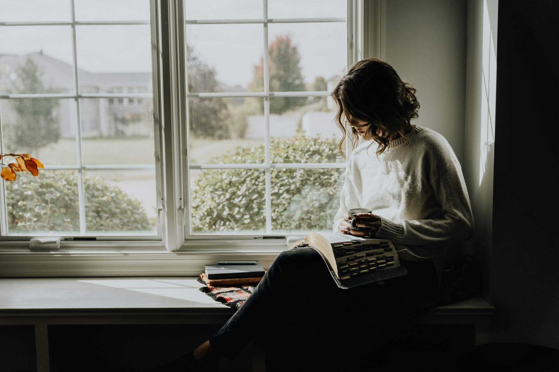 A person sits by a window reading a book, holding a mug, with several other books stacked nearby—capturing the peaceful focus of mums studying or returning to study. Daylight streams through the window.