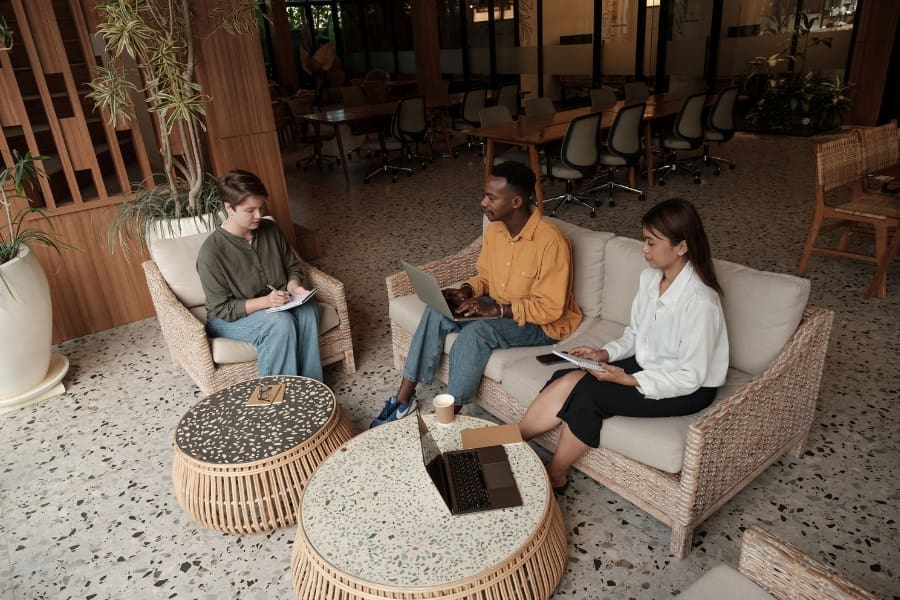 Three people are sitting in a modern lounge area with laptops and notebooks, engaging in an essential discussion about new business strategies.
