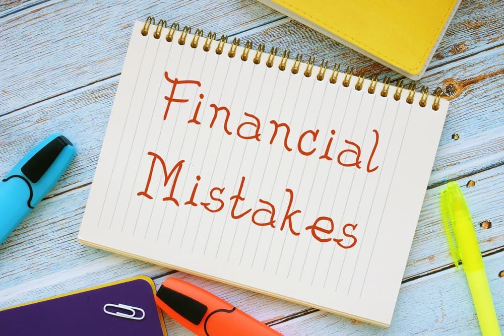 A notebook open to a page with "Financial Mistakes" written in red text, surrounded by highlighters and folders on a wooden surface.