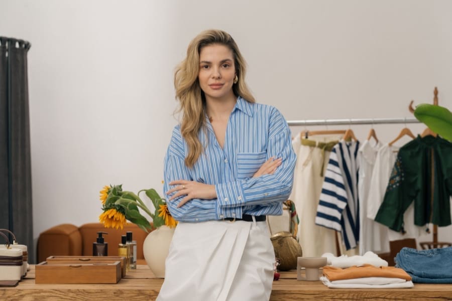A woman with long blonde hair stands with arms crossed in front of a table with sunflowers, clothing, and accessories—a vibrant scene that celebrates mum-run businesses and their drive to thrive. A clothing rack with shirts and jackets is in the background.