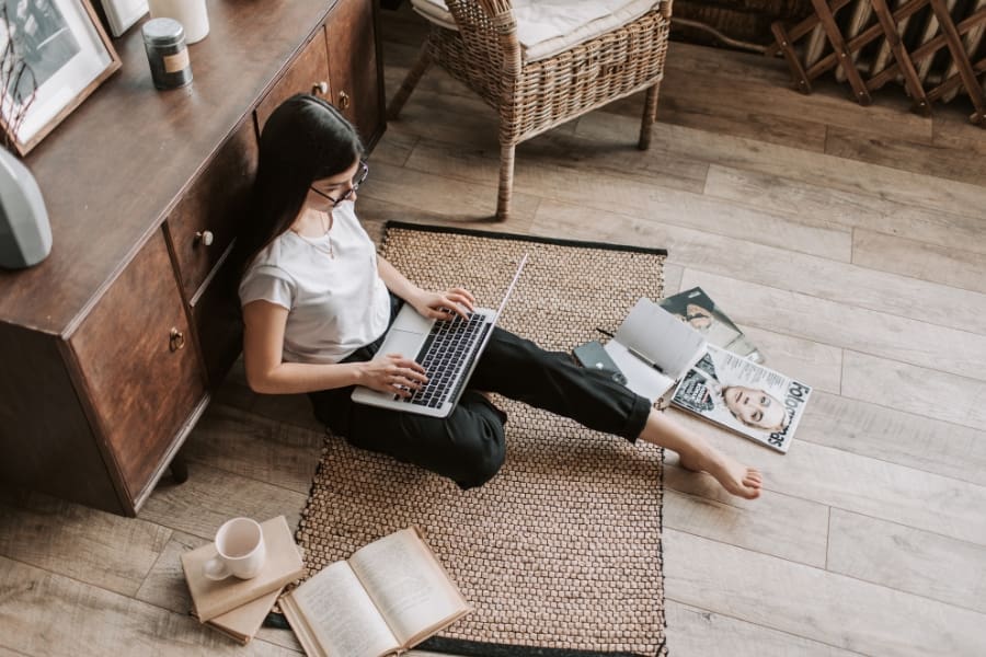 A mum blogger sits on a rug on a wooden floor, working on her laptop, surrounded by books, magazines, and a cup—balancing it all as she works to build authority and avoid burnout.