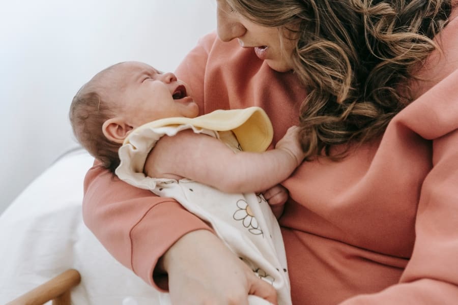 A woman holds a crying infant in her arms, comforting them indoors while anxiously checking for signs of infant choking or mucus, following a parent guide for reassurance.